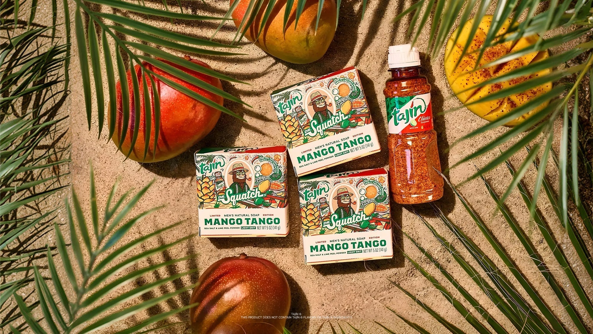 Three bars of Tajín Mango Tango soap and a bottle of Tajín seasoning arranged on sand with tropical palm leaves and mangoes.