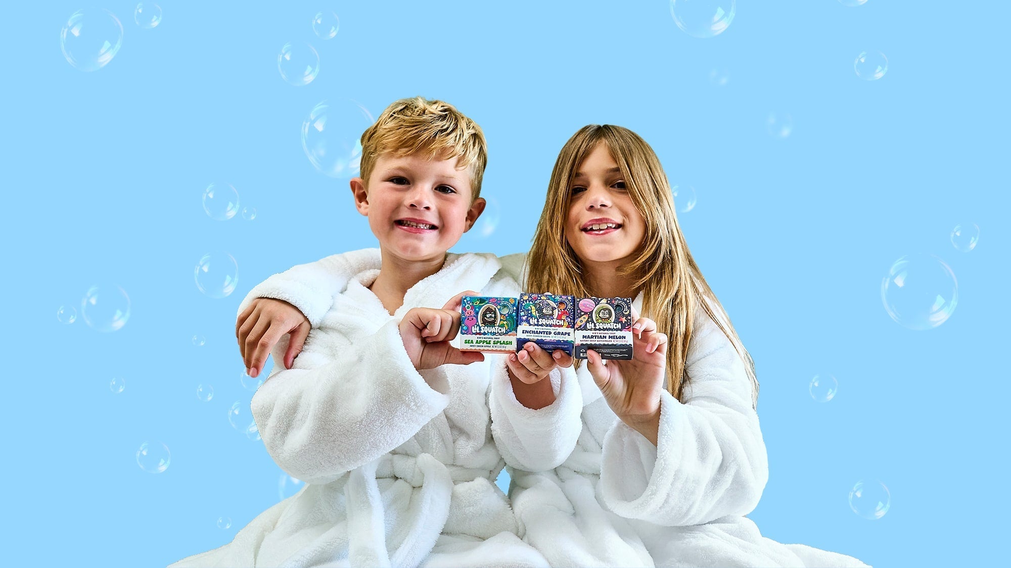 Two children in white robes holding three colorful Lil Squatch product boxes against a blue background with bubbles.
